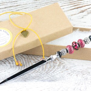 May include: A decorative letter opener with a black blade and a beaded handle. The handle features pink leopard print beads, silver beads, and black accents. The opener is presented in a cardboard box with a yellow cord and a heart charm.