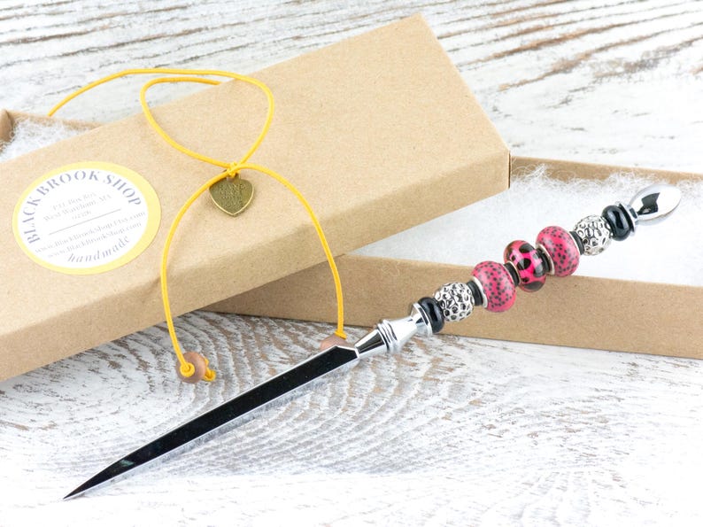 May include: A decorative letter opener with a black blade and a beaded handle. The handle features pink leopard print beads, silver beads, and black accents. The opener is presented in a cardboard box with a yellow cord and a heart charm.