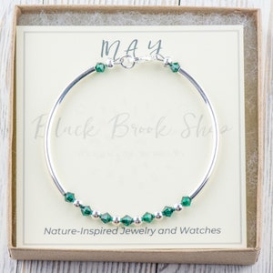 May include: A silver bangle bracelet with a row of emerald green crystals and the word "MAY" on a white card in a box.