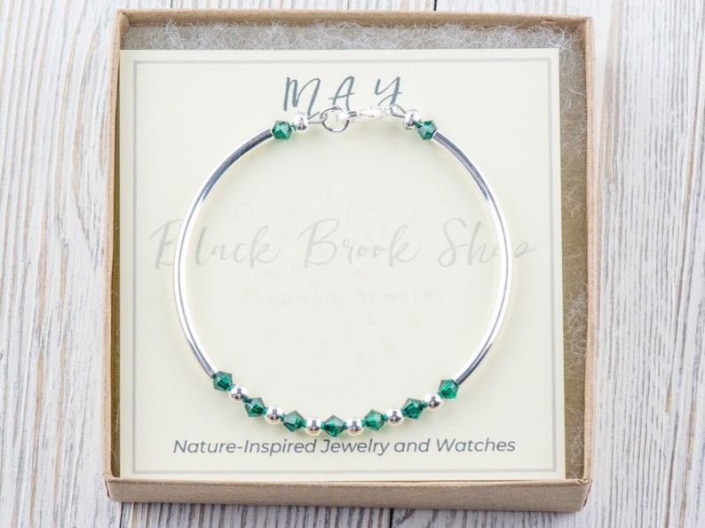 May include: A silver bangle bracelet with a row of emerald green crystals and the word "MAY" on a white card in a box.