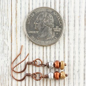 May include: Copper-colored earrings with silver, bronze, and gold-colored beads are displayed next to a U.S. quarter. The earrings have a simple, elegant design with a hook closure. The background is a white-painted wooden surface.
