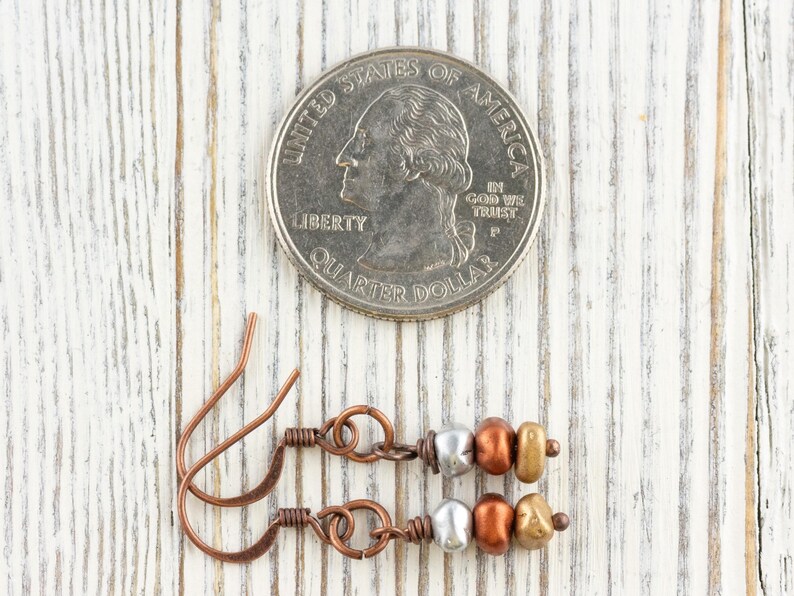 May include: Copper-colored earrings with silver, bronze, and gold-colored beads are displayed next to a U.S. quarter. The earrings have a simple, elegant design with a hook closure. The background is a white-painted wooden surface.
