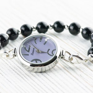 May include: A silver watch with a purple face and black numbers. The watch is attached to a black beaded bracelet with a silver clasp.