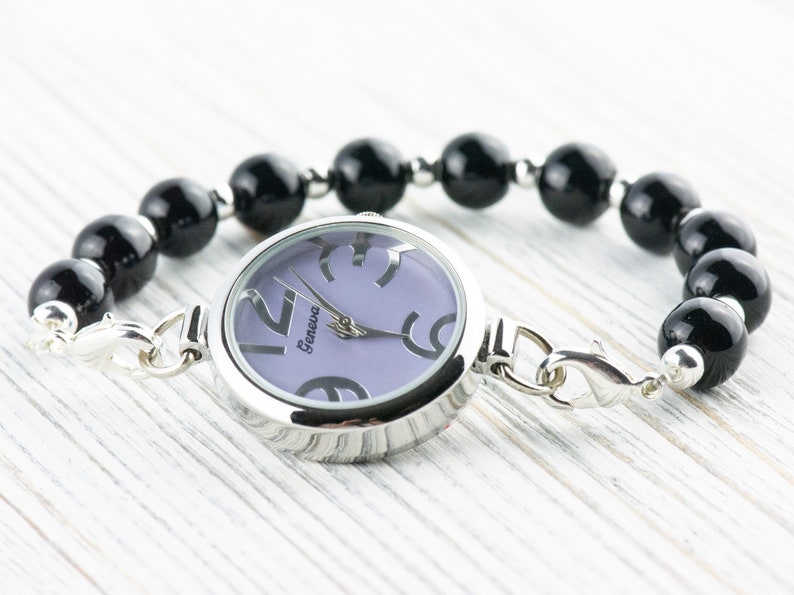 May include: A silver watch with a purple face and black numbers. The watch is attached to a black beaded bracelet with a silver clasp.