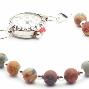 May include: A silver-toned watch bracelet with a white watch face and a red crown. The bracelet features a series of multi-colored, spherical beads. The beads are connected by small silver-colored metal beads.