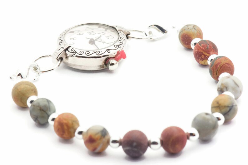 May include: A silver-toned watch bracelet with a white watch face and a red crown. The bracelet features a series of multi-colored, spherical beads. The beads are connected by small silver-colored metal beads.