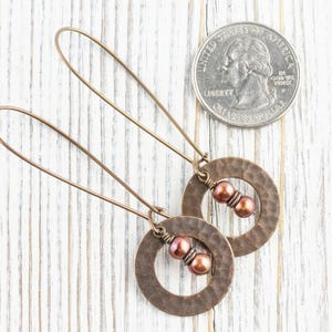 May include: A pair of copper-colored earrings with a hammered texture. Each earring features a circular design with two reddish-brown beads and a long, thin wire for the ear. A US quarter is next to the earrings for size comparison.