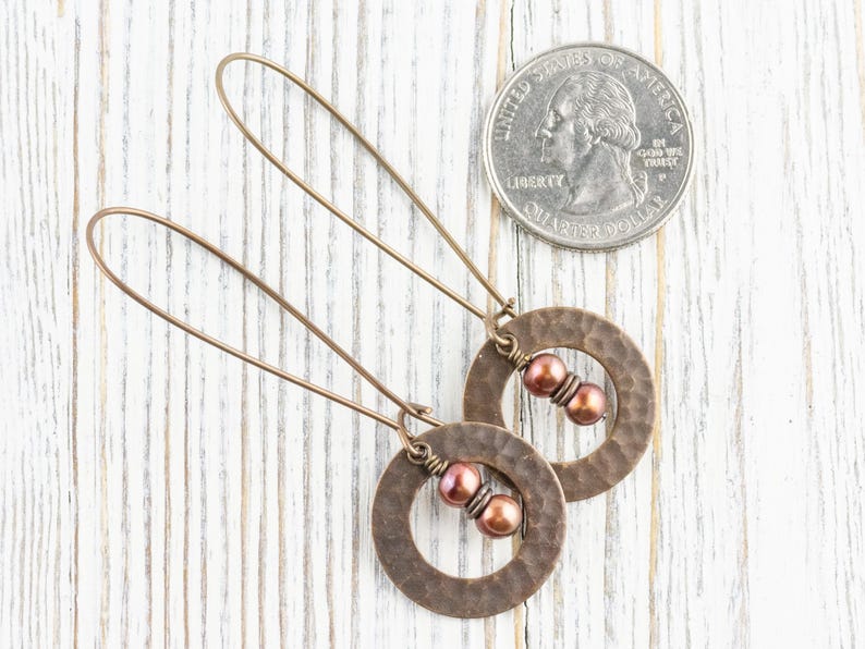 May include: A pair of copper-colored earrings with a hammered texture. Each earring features a circular design with two reddish-brown beads and a long, thin wire for the ear. A US quarter is next to the earrings for size comparison.