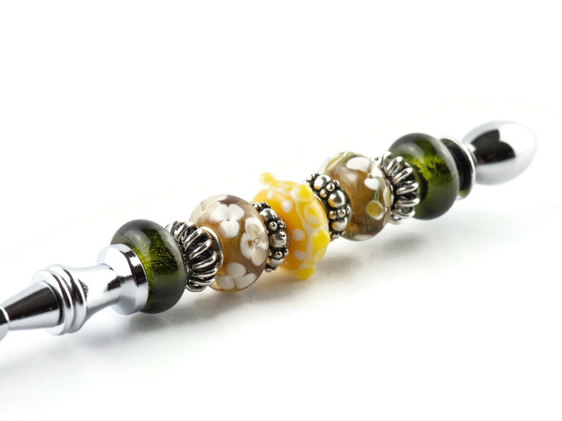 K&ouml;nnte beinhalten: Ein silberner und verchromter Stift mit einem Design aus gr&uuml;nen, gelben und braunen Glasperlen. Die Perlen haben wei&szlig;e Blumenmuster.