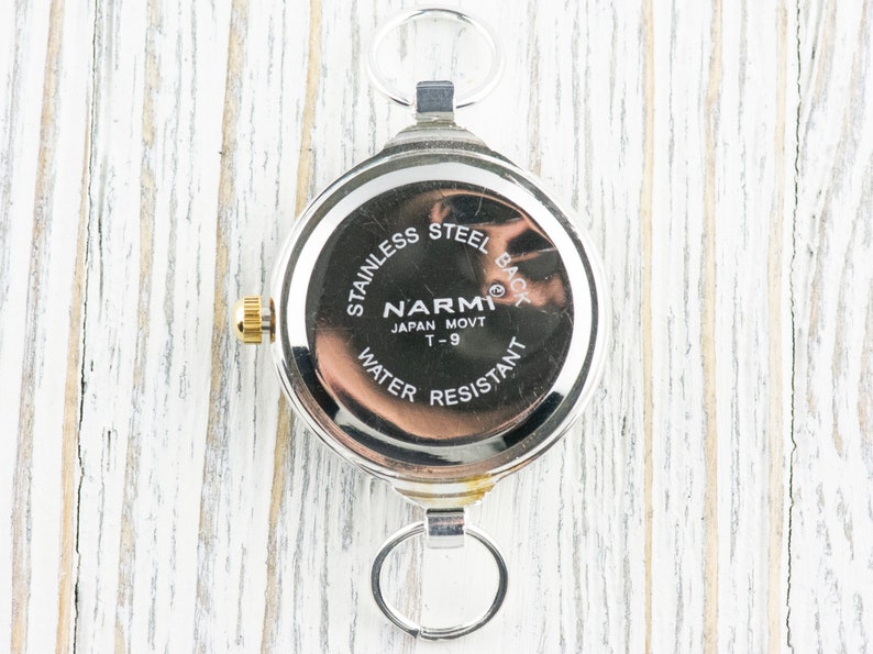 Pu&ograve; includere: Un orologio da tasca argentato con corona dorata e retro in acciaio inossidabile. Presenta anelli per il fissaggio in alto e in basso. Il retro reca la scritta "STAINLESS STEEL BACK", "NARMI JAPAN MOVT T-9" e "WATER RESISTANT".