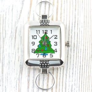 Puede incluir: Reloj de llavero cuadrado plateado con esfera blanca y dise&ntilde;o de &aacute;rbol de Navidad verde. El reloj tiene n&uacute;meros y manecillas negras, y una peque&ntilde;a corona. Est&aacute; sujeto a un llavero plateado. Accesorio festivo.
