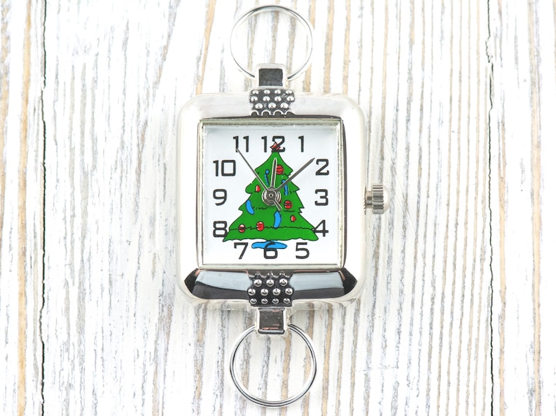 Puede incluir: Reloj de llavero cuadrado plateado con esfera blanca y dise&ntilde;o de &aacute;rbol de Navidad verde. El reloj tiene n&uacute;meros y manecillas negras, y una peque&ntilde;a corona. Est&aacute; sujeto a un llavero plateado. Accesorio festivo.
