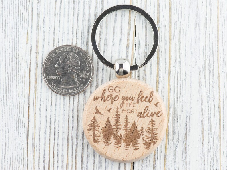 May include: Wooden keychain with a brown background and the text "Go where you feel the most alive" engraved on it. The keychain features a silver key ring and a design of trees.
