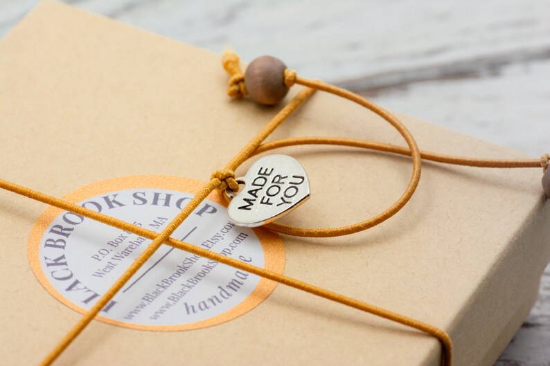 May include: A brown cardboard box with a yellow string tied around it. The string has a silver heart charm with the words "MADE FOR YOU" on it. The box has a round sticker with the text "BLACK BROOK SHOP" and contact information.