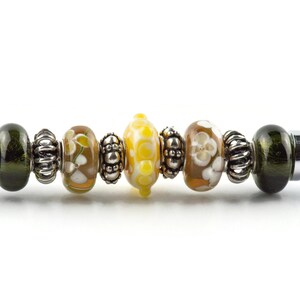 K&ouml;nnte beinhalten: Eine Nahaufnahme eines silbernen Charm-Armbands mit verschiedenen bunten Glasperlen. Die Perlen sind gr&uuml;n, gelb, braun und wei&szlig; mit Blumenmustern. Das Armband ist aus Metall und hat einen silbernen Verschluss.
