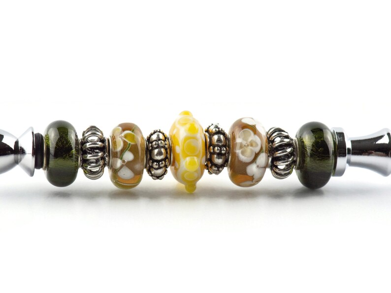 K&ouml;nnte beinhalten: Eine Nahaufnahme eines silbernen Charm-Armbands mit verschiedenen bunten Glasperlen. Die Perlen sind gr&uuml;n, gelb, braun und wei&szlig; mit Blumenmustern. Das Armband ist aus Metall und hat einen silbernen Verschluss.