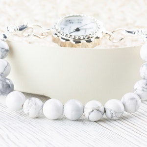 Peut inclure: Un bracelet en perles blanches et grises avec une montre argent&eacute;e attach&eacute;e. La montre a un cadran blanc avec des chiffres et des aiguilles noirs.