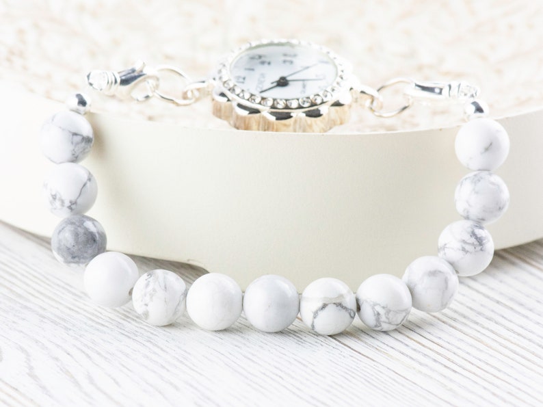 Peut inclure: Un bracelet en perles blanches et grises avec une montre argent&eacute;e attach&eacute;e. La montre a un cadran blanc avec des chiffres et des aiguilles noirs.
