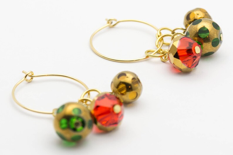 Puede incluir: Aretes de aro de oro con colgantes. Los colgantes son cuentas de vidrio rojo, verde y dorado con un patr&oacute;n moteado.