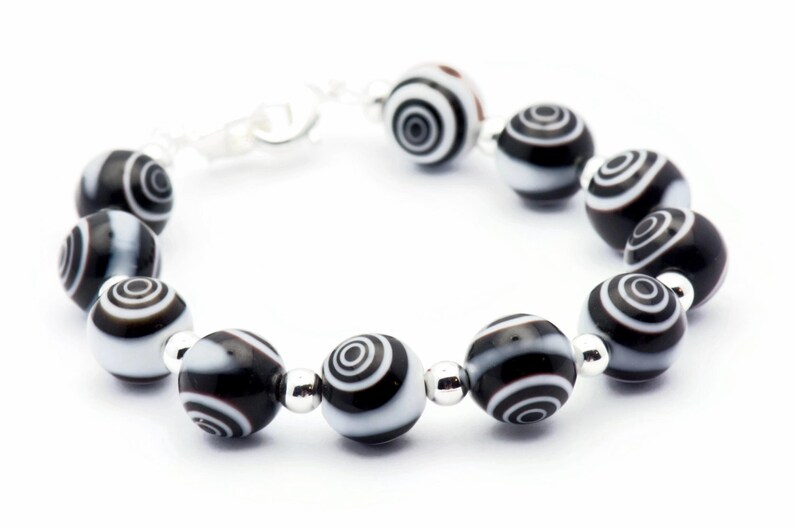 K&ouml;nnte beinhalten: Ein schwarz-wei&szlig;es Armband mit einem silbernen Verschluss. Das Armband besteht aus runden Perlen mit einem konzentrischen Kreis-Muster.