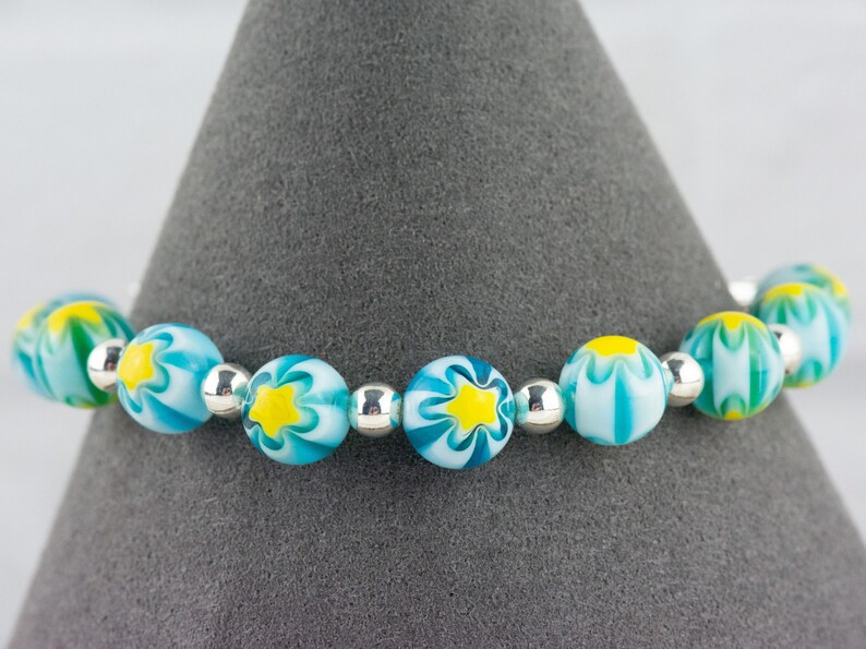 Peut inclure: Un bracelet compos&eacute; de perles de verre bleues et blanches avec des motifs de fleurs jaunes et des perles d'espacement argent&eacute;es.