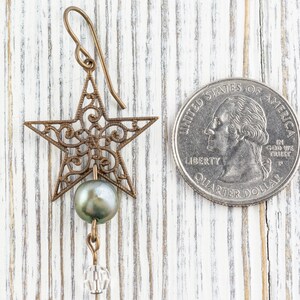 May include: A bronze-colored star-shaped earring with intricate filigree design, a gray pearl, and a clear crystal bead. The earring is displayed next to a U.S. quarter coin for size comparison. The background is a white wooden surface.