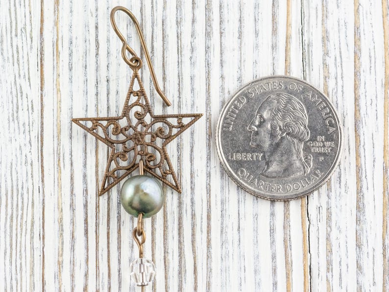 May include: A bronze-colored star-shaped earring with intricate filigree design, a gray pearl, and a clear crystal bead. The earring is displayed next to a U.S. quarter coin for size comparison. The background is a white wooden surface.