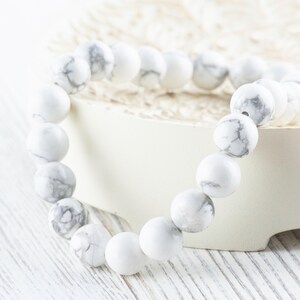 Pode incluir: Pulseira de contas de howlite branca com veios cinzentos. A pulseira &eacute; feita de contas redondas e est&aacute; sobre uma superf&iacute;cie branca.