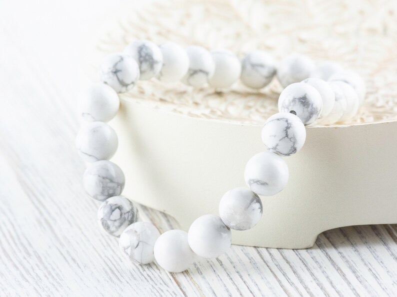 Pode incluir: Pulseira de contas de howlite branca com veios cinzentos. A pulseira &eacute; feita de contas redondas e est&aacute; sobre uma superf&iacute;cie branca.