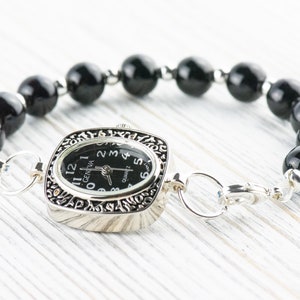 K&ouml;nnte beinhalten: Ein schwarzes Perlenarmband mit einer silbernen Uhr. Das Zifferblatt der Uhr ist quadratisch und hat schwarze Zeiger und Zahlen. Das Zifferblatt der Uhr zeigt "GENEVA" und "QUARTZ".