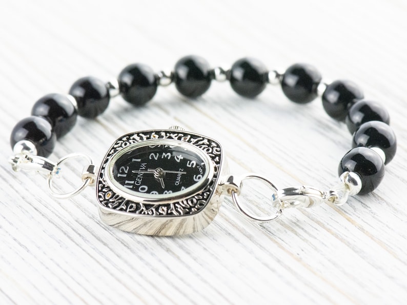K&ouml;nnte beinhalten: Ein schwarzes Perlenarmband mit einer silbernen Uhr. Das Zifferblatt der Uhr ist quadratisch und hat schwarze Zeiger und Zahlen. Das Zifferblatt der Uhr zeigt "GENEVA" und "QUARTZ".