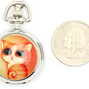 Op de afbeelding: Zilveren zakhorloge met een ketting, met een cartoonkat met oranje en witte vacht en grote ogen. De wijzerplaat van het horloge is oranje en geel.