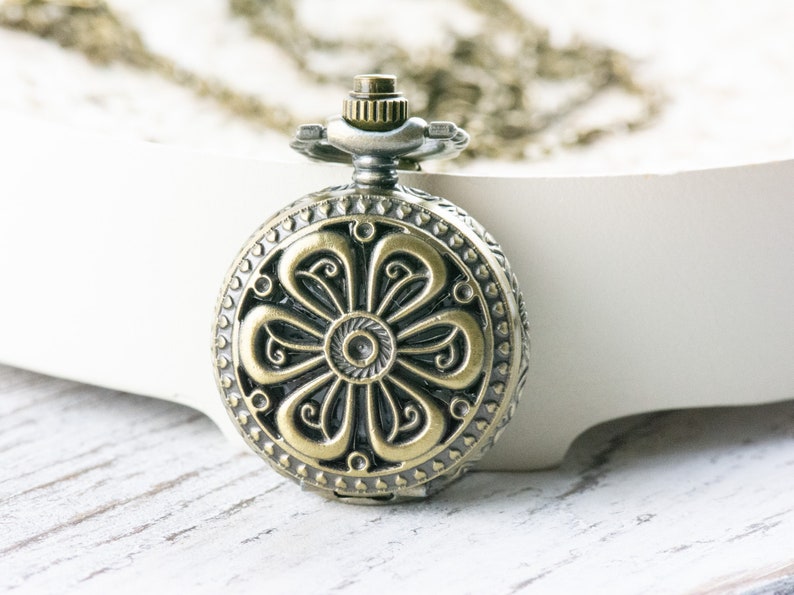 May include: A gold-toned pocket watch with a floral design on the face. The watch is attached to a chain.