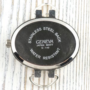 May include: A close-up of a black Geneva watch back with silver detailing. The oval-shaped watch back has the text "STAINLESS STEEL BACK", "GENEVA JAPAN MOVT S-140", and "WATER RESISTANT". It features silver loops for a strap.