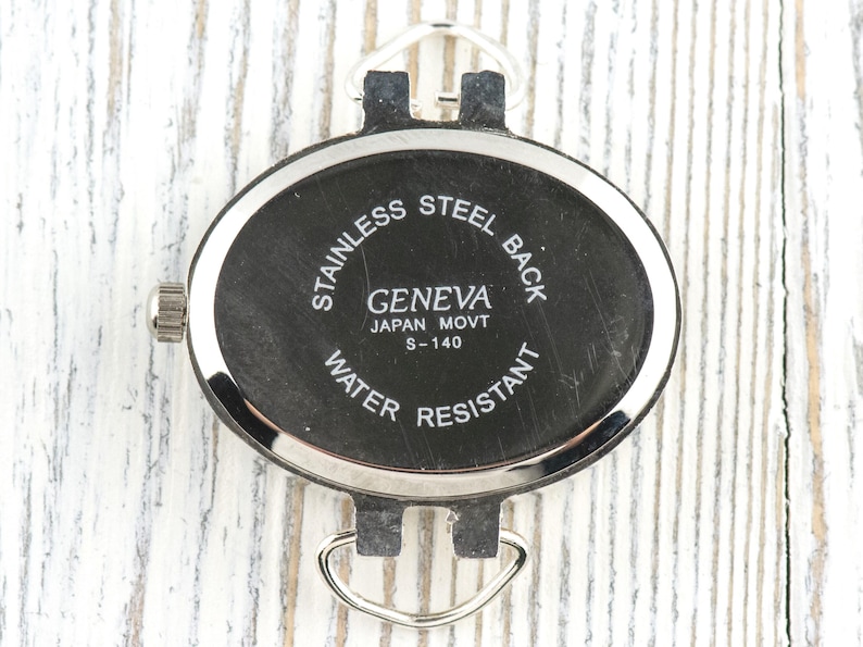 May include: A close-up of a black Geneva watch back with silver detailing. The oval-shaped watch back has the text "STAINLESS STEEL BACK", "GENEVA JAPAN MOVT S-140", and "WATER RESISTANT". It features silver loops for a strap.