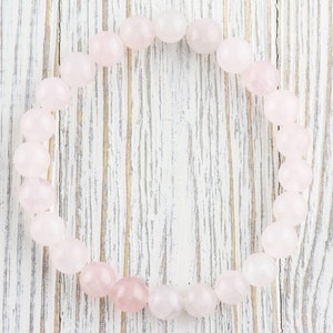 May include: A pink rose quartz bracelet made of smooth, round beads. The bracelet is laid out on a white wooden surface.