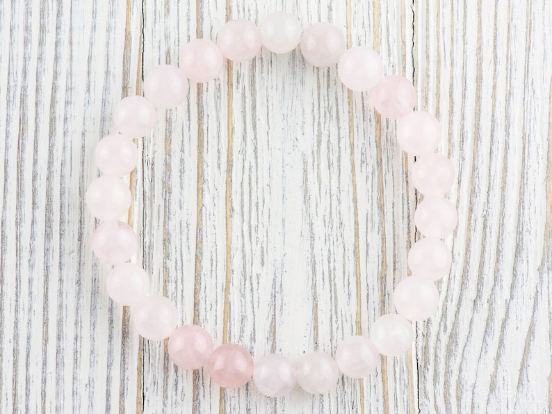 May include: A pink rose quartz bracelet made of smooth, round beads. The bracelet is laid out on a white wooden surface.