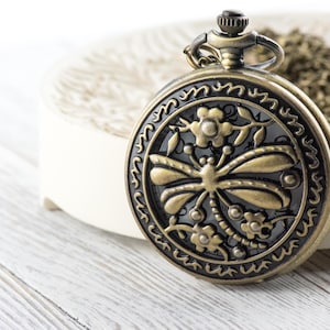 May include: Antique bronze pocket watch with a dragonfly design. The watch features a detailed floral and insect motif on the face. The watch is round and has a chain. The watch is on a white surface.