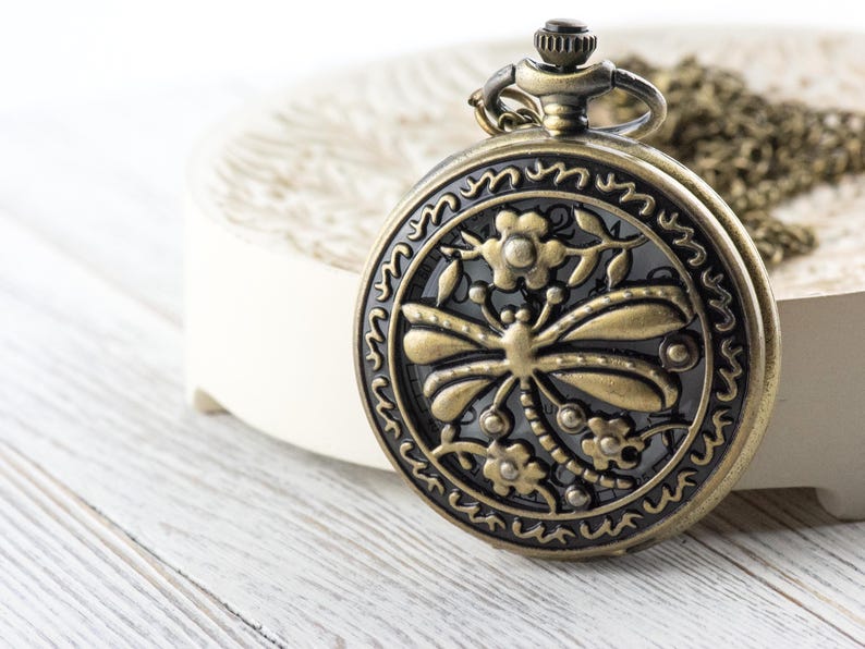 May include: Antique bronze pocket watch with a dragonfly design. The watch features a detailed floral and insect motif on the face. The watch is round and has a chain. The watch is on a white surface.