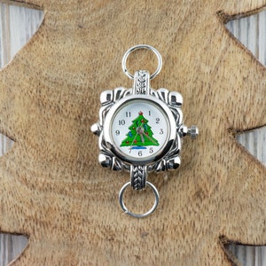 May include: A silver watch with a white face and a green Christmas tree design. The watch has a silver band with a decorative clasp.