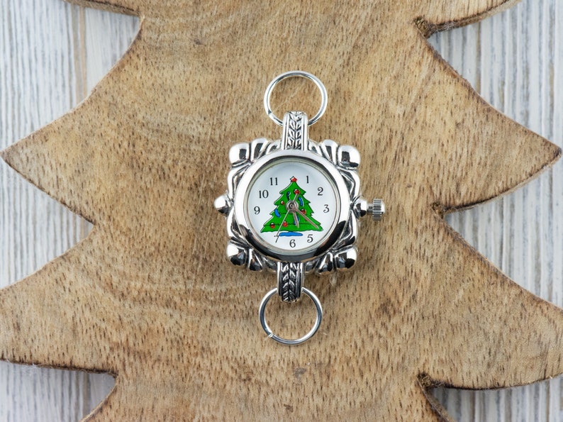 May include: A silver watch with a white face and a green Christmas tree design. The watch has a silver band with a decorative clasp.