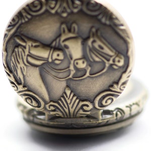 Puede incluir: Un reloj de bolsillo dorado con un dise&ntilde;o ornamentado que presenta dos caballos. La esfera del reloj est&aacute; oculta detr&aacute;s de la tapa.