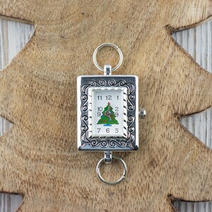 Op de afbeelding: Een zilveren zakhorloge met een kerstboomontwerp op de wijzerplaat. Het horloge heeft een decoratieve rand en een lus om op te hangen.