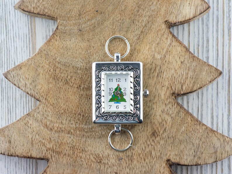Op de afbeelding: Een zilveren zakhorloge met een kerstboomontwerp op de wijzerplaat. Het horloge heeft een decoratieve rand en een lus om op te hangen.