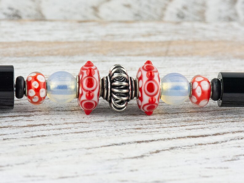 Puede incluir: Primer plano de una pulsera con cuentas de vidrio rojas y blancas, una cuenta espaciadora plateada y tapas finales negras.