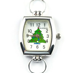 May include: Silver-toned Christmas-themed watch with a white face featuring a green Christmas tree design. The watch has a rectangular face with rounded edges, a crown, and two circular loops for attachment.