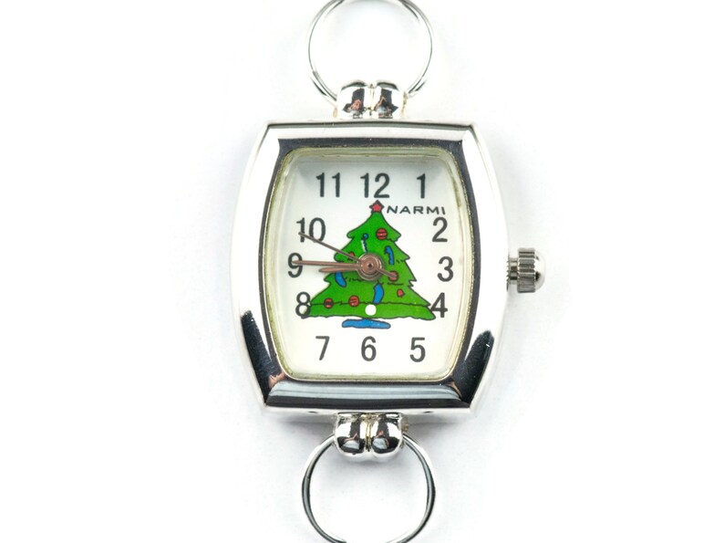 May include: Silver-toned Christmas-themed watch with a white face featuring a green Christmas tree design. The watch has a rectangular face with rounded edges, a crown, and two circular loops for attachment.