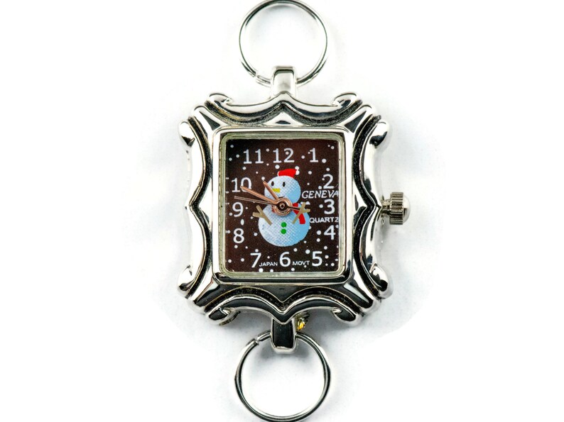May include: A silver pocket watch with a brown face and a snowman design. The watch face reads "GENEVA QUARTZ" and "JAPAN MVT".