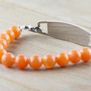 Op de afbeelding: Een zilveren armband met een rechthoekig label en oranje kralen. Het label is leeg en kan worden gegraveerd.