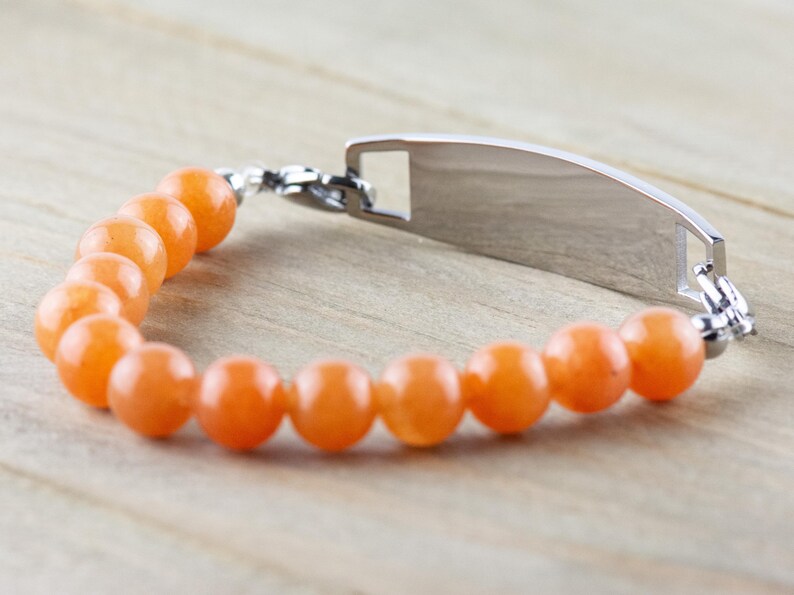 Op de afbeelding: Een zilveren armband met een rechthoekig label en oranje kralen. Het label is leeg en kan worden gegraveerd.